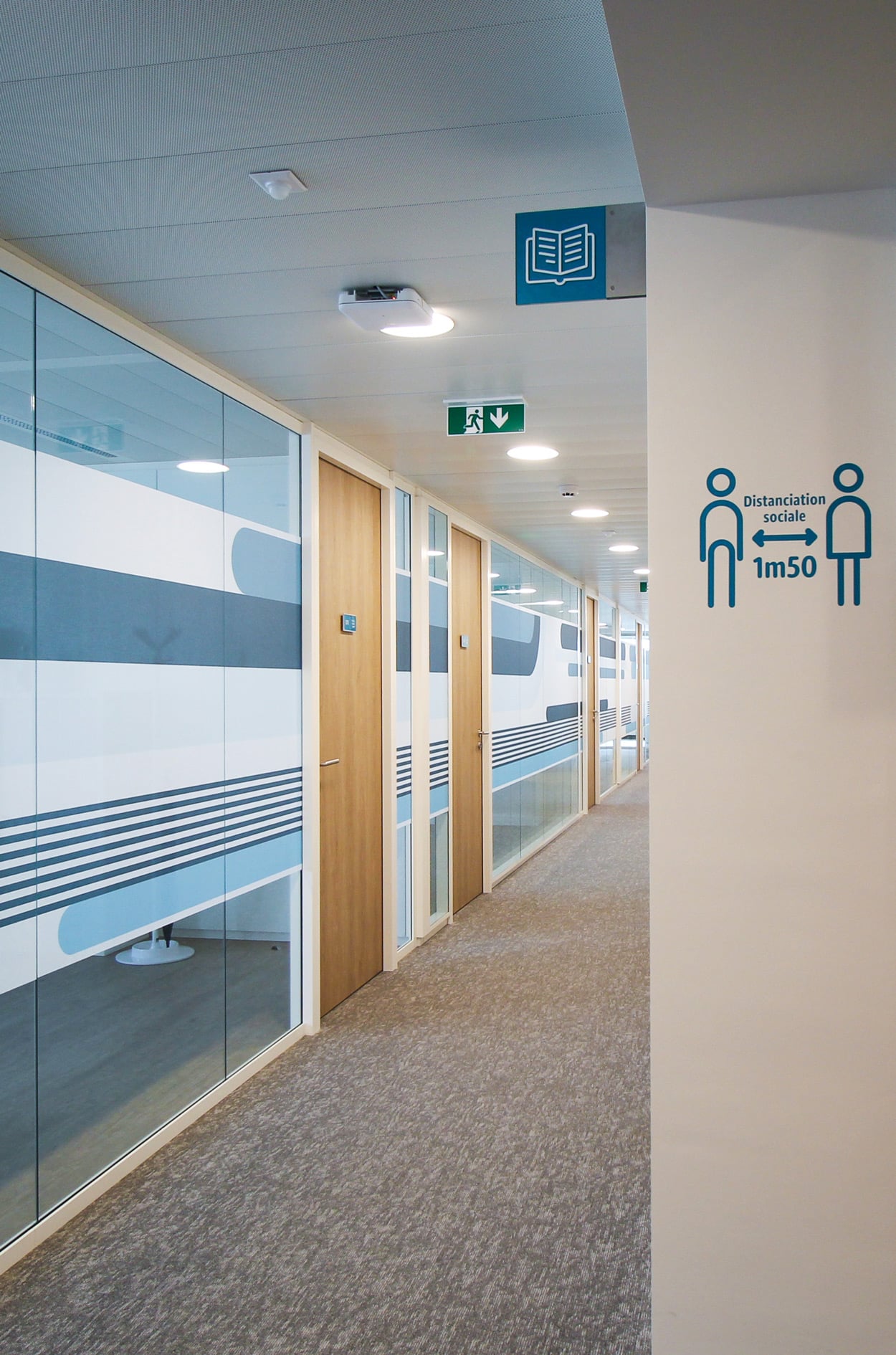 Vue d'ambiance d'un couloir avec plaques de port Skwizmi Marcal sur portes en bois, drapeau directionnel bibliotheque et stickers distanciation sociale et habillage décoratif sur cloisons vitrées en tissus autocollant design (Twodesigners) des bandes dans les tons de bleu